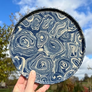 Op de afbeelding: Een ronde keramische plaat met een uniek marmerpatroon. De plaat heeft een wervelend patroon van marineblauw en crèmekleur. De plaat wordt omhoog gehouden tegen een achtergrond van een blauwe lucht en groen gebladerte.