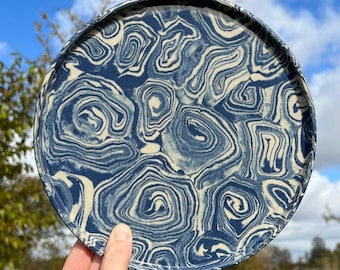 May include: A circular ceramic plate with a distinctive marbled pattern. The plate displays a swirling design in navy blue and cream. The plate is held up against a backdrop of a blue sky and green foliage.
