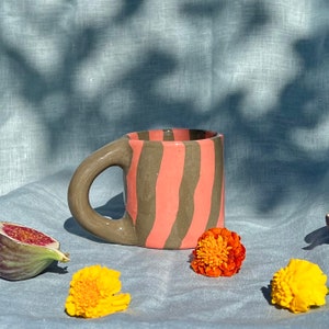 May include: A ceramic mug with a large handle, featuring vertical stripes in coral and brown. The mug is surrounded by figs and yellow and orange flowers, all set on a light blue cloth.