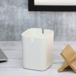 May include: A white square-shaped desk organizer with a small slot on the front, next to a wooden phone stand. The organizer and stand are on a white wooden surface.