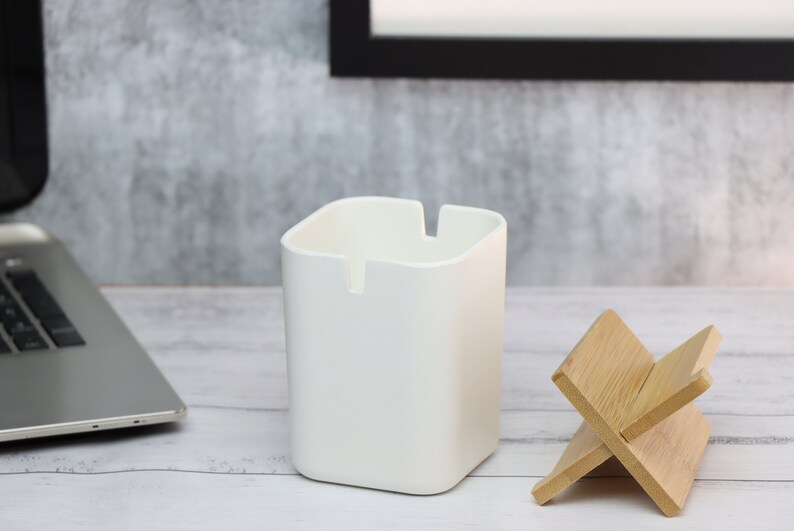 May include: A white square-shaped desk organizer with a small slot on the front, next to a wooden phone stand. The organizer and stand are on a white wooden surface.