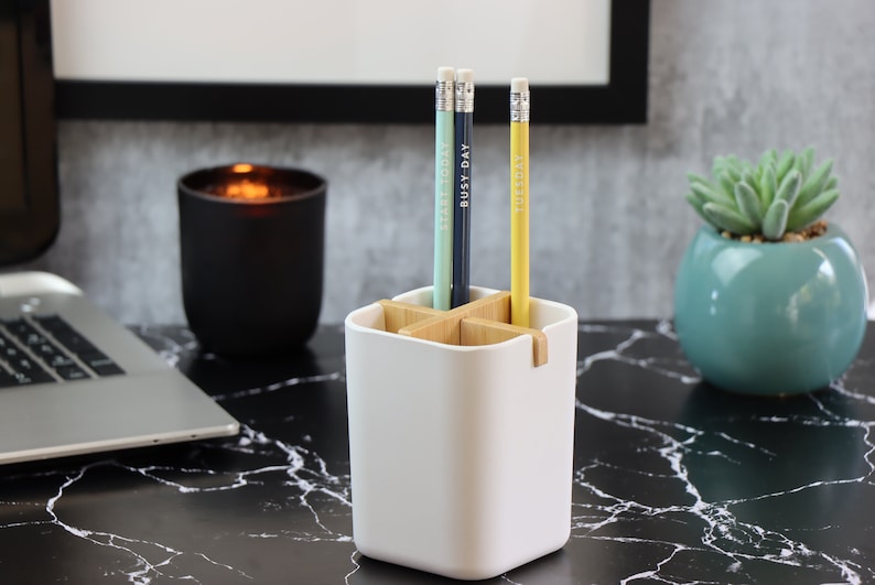May include: White ceramic desk organizer with a wooden divider. Three pencils are in the organizer, two are blue and one is yellow. The pencils have the words "Busy Day" and "Tuesday" written on them.