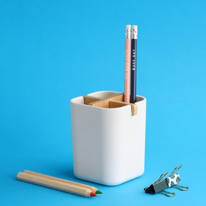 May include: White ceramic desk organizer with four compartments, two of which are filled with pencils. The pencils are pink and blue with the words "Good Days" and "Busy Day" written on them. A wooden pencil and a silver and blue patterned paper clip are on a blue surface.