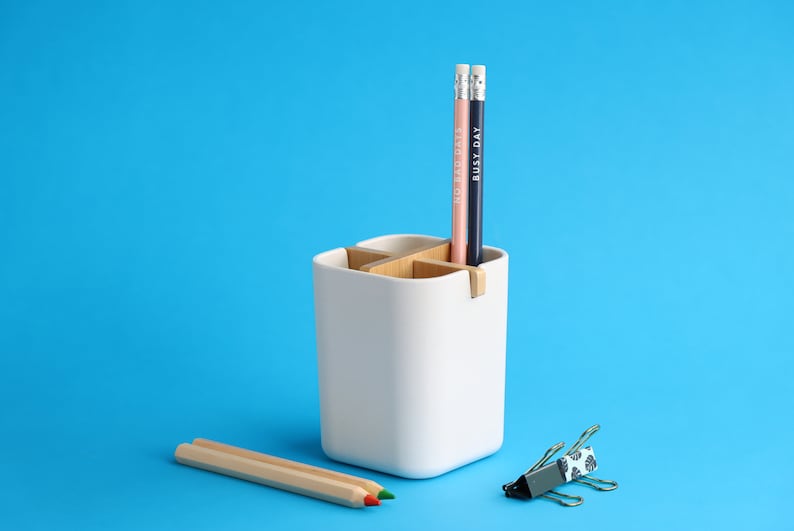 May include: White ceramic desk organizer with four compartments, two of which are filled with pencils. The pencils are pink and blue with the words "Good Days" and "Busy Day" written on them. A wooden pencil and a silver and blue patterned paper clip are on a blue surface.