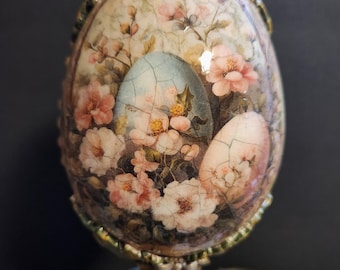 Op de afbeelding: Een decoratief paasei met een bloemenmotief en een houten standaard. Het ei is beschilderd met roze en witte bloemen op een blauwe achtergrond. De standaard is van hout en heeft een bruine afwerking.