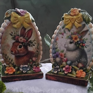 以下が含まれることがあります： イースターをテーマにした2つの装飾品。それぞれ、花の冠をかぶったウサギの肖像画が、卵型のフレームに収められています。フレームは黄色のリボンと花のアクセントで飾られ、木製の台座の上に置かれています。