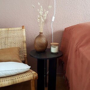 以下が含まれることがあります： 黒のサイドテーブルに、質感のある茶色の花瓶とガラスのジャーに入った白いキャンドルが置かれています。テーブルの隣には、白いクッションと茶色のクッションを置いた籐製の椅子があります。壁は薄いピンク色です。