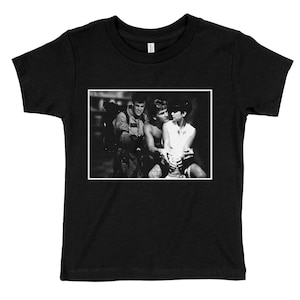 Puede incluir: Foto en blanco y negro de tres personas. Dos hombres y una mujer están de pie juntos. Los hombres visten ropa informal y la mujer lleva una camisa blanca. La foto está impresa en una camiseta negra.