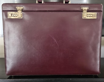May include: A rectangular burgundy hard-shell briefcase featuring a smooth finish and a top handle. The front is secured with two gold-toned metal combination locks, one on each side, which include three-digit rotating number dials.