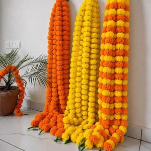 May include: Three long decorative garlands made of artificial marigold flowers hanging vertically. One garland is solid orange, one is solid yellow, and the third features an alternating pattern of orange and yellow flower clusters.