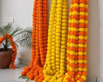 Könnte beinhalten: Drei lange dekorative Girlanden aus künstlichen Ringelblumen, die vertikal hängen. Eine Girlande ist einfarbig orange, eine einfarbig gelb und die dritte zeigt ein abwechselndes Muster aus orangefarbenen und gelben Blütenbüscheln.