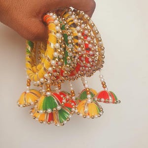 May include: A set of vibrant bangles featuring yellow, red, and green threads, embellished with gold and white beads. The bangles have decorative bell-shaped charms hanging from them. The design is intricate and eye-catching, suitable for special occasions.