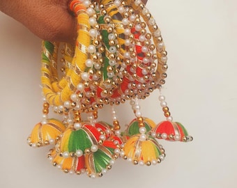 Può includere: Una collezione di braccialetti colorati con dettagli intricati. I braccialetti sono adornati con fili gialli, rossi e verdi, accentuati da perline dorate e bianche. Ciondoli decorativi a forma di campana pendono dai braccialetti.