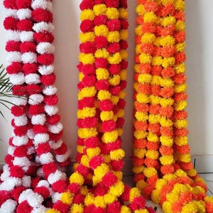 May include: Three strands of decorative flower garlands. One is red and white, the second is red, yellow, and the third is orange and yellow. The garlands are made of small, round, pom-pom-like flowers, and are hanging vertically.