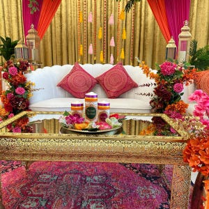 Puede incluir: Una mesa de café de espejo dorado con detalles intrincados se encuentra sobre una alfombra rosa y morada. La mesa está decorada con tres velas blancas, flores rosas y naranjas, y un plato pequeño. La mesa está frente a un sofá blanco con almohadas rosas y doradas. El fondo está decorado con cortinas rosas y naranjas y borlas doradas.