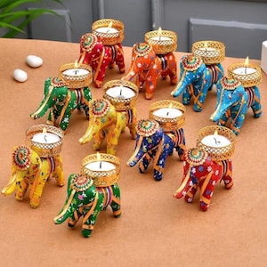 May include: A collection of colorful elephant-shaped candle holders. Each elephant is painted in a different color, including red, green, blue, yellow, and orange, and has a gold-colored metal basket on its back to hold a lit candle. The candle holders are arranged on a brown surface.