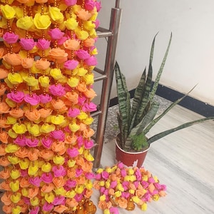 May include: A colorful floral garland with yellow, pink, and orange roses. The garland is hanging, and a small pile of matching flowers rests on the floor. Gold-colored bells hang at the bottom of the garland.
