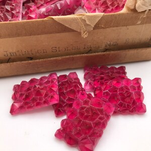 Puede incluir: Una caja de piedras de imitación de vidrio rosa, etiquetada como "Imitation Stones Fac No 814". La caja está abierta y varias piedras están esparcidas sobre una superficie blanca.