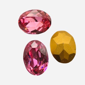 Puede incluir: Tres gemas ovales facetadas. Dos son de un rosa vibrante y una es de color oro metálico. Las gemas se muestran sobre un fondo blanco, mostrando su talla y color.