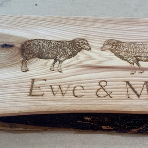 Puede incluir: Una tabla de cortar de madera con un acabado de veta de madera natural. La tabla tiene un asa y está decorada con un diseño de dos ovejas quemado en madera y el texto "Ewe & Me".