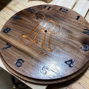 May include: A wooden clock face with black Roman numeral hour markers and a black number 4. The clock face has a large, engraved letter "R" in the center with the text "EST. JULY 4, 2020" underneath.