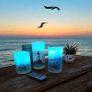 以下が含まれることがあります： 灯台と帆船の航海デザインが施された、3つの青いLEDキャンドル。リモコンがキャンドルの前にあります。装飾的なポットに入った小さな多肉植物も存在します。背景には海上の夕焼けが写っています。