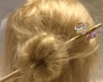 Puede incluir: Un palo de pelo de madera con un diseño floral decorativo con flores y perlas iridiscentes azules, moradas y blancas. El palo está insertado en un moño rubio.