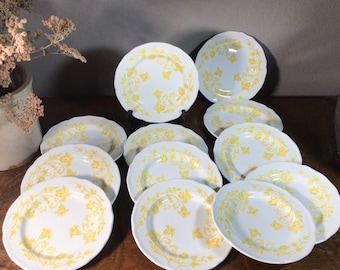 May include: A set of 12 white ceramic plates with a yellow floral design. The plates are arranged in a stack on a wooden surface.
