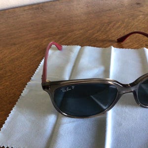 Puede incluir: Gafas de sol Ray-Ban con montura marr&oacute;n y lentes oscuras. Las patillas son de color rosa rojizo. Las gafas de sol est&aacute;n sobre un pa&ntilde;o de limpieza blanco.