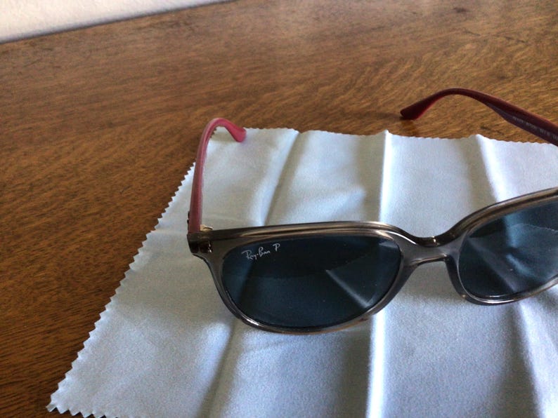 Puede incluir: Gafas de sol Ray-Ban con montura marr&oacute;n y lentes oscuras. Las patillas son de color rosa rojizo. Las gafas de sol est&aacute;n sobre un pa&ntilde;o de limpieza blanco.