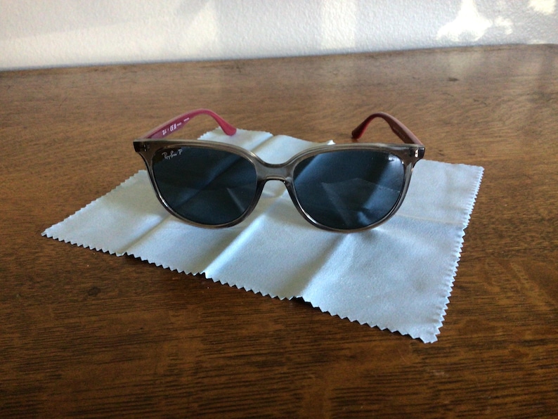 Puede incluir: Gafas de sol con montura y lentes oscuras, sobre un pa&ntilde;o de limpieza blanco. Las patillas son de color rosa rojizo. Las gafas de sol est&aacute;n sobre una superficie de madera.