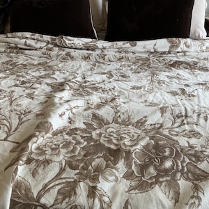 May include: A white bedspread featuring a brown floral pattern with detailed illustrations of blooming flowers and leaves. The fabric is draped across a bed, with two dark brown pillows visible at the top edge of the frame, providing a contrast.