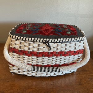 Pode incluir: Uma cesta tecida com tampa articulada e alça. A cesta apresenta um padrão branco e vermelho com uma tampa cinza adornada com um desenho floral em vermelho, azul e verde. A alça é um material espesso branco semelhante a uma corda.