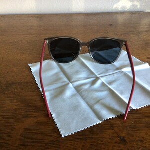 Puede incluir: Gafas de sol con montura y lentes oscuras, y patillas rojas. Se encuentran sobre un pa&ntilde;o de limpieza blanco, sobre una mesa de madera. El dise&ntilde;o es moderno y elegante.