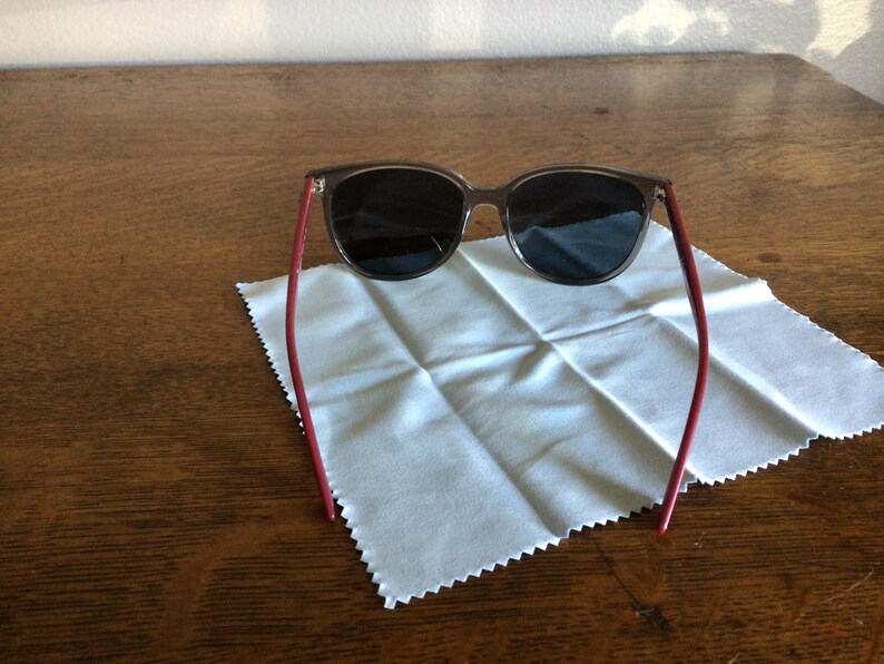 Puede incluir: Gafas de sol con montura y lentes oscuras, y patillas rojas. Se encuentran sobre un pa&ntilde;o de limpieza blanco, sobre una mesa de madera. El dise&ntilde;o es moderno y elegante.