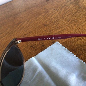 Puede incluir: Gafas de sol con lentes oscuras y una patilla de color burdeos. La patilla muestra el logotipo de Ray-Ban, la marca CE y la palabra "POLARIZED". Se ve un pa&ntilde;o de limpieza azul claro.