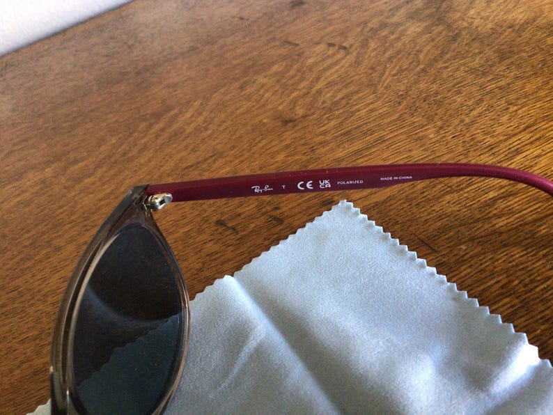 Puede incluir: Gafas de sol con lentes oscuras y una patilla de color burdeos. La patilla muestra el logotipo de Ray-Ban, la marca CE y la palabra "POLARIZED". Se ve un pa&ntilde;o de limpieza azul claro.