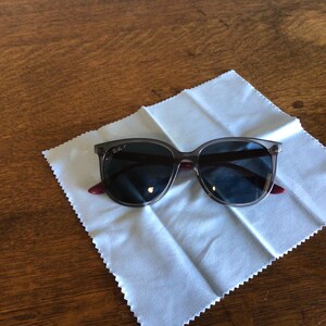 Puede incluir: Gafas de sol con lentes oscuras y montura gris transl&uacute;cida sobre un pa&ntilde;o de limpieza blanco. Las patillas tienen un acento rojo. Las gafas de sol est&aacute;n sobre una superficie de madera. Un accesorio de moda.