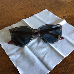 Puede incluir: Gafas de sol con lentes oscuras y montura transl&uacute;cida sobre un pa&ntilde;o de limpieza blanco. Las patillas tienen un acento burdeos. El logotipo de Ray-Ban es visible en la patilla izquierda. Las gafas de sol est&aacute;n sobre una superficie de madera.