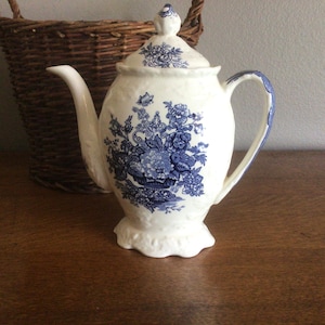 Op de afbeelding: Een witte keramische theepot met een blauw bloemmotief. De theepot heeft een gebogen tuit, een handvat en een deksel met een kleine knop. Het bloemmotief is gedetailleerd en bedekt het grootste deel van het oppervlak van de theepot.