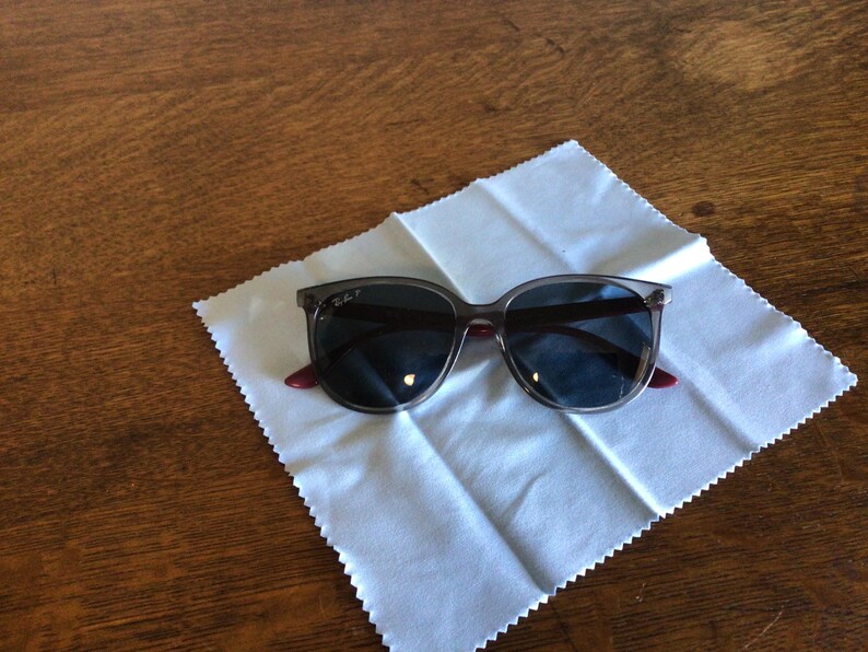 Puede incluir: Gafas de sol con lentes oscuras y montura gris transl&uacute;cida, sobre un pa&ntilde;o de limpieza blanco. Las patillas tienen un acento rojo. Las gafas de sol est&aacute;n sobre una superficie de madera.
