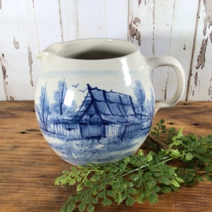 May include: A blue and white ceramic pitcher with a hand-painted scene of a house and trees. The pitcher has a rounded body and a handle.