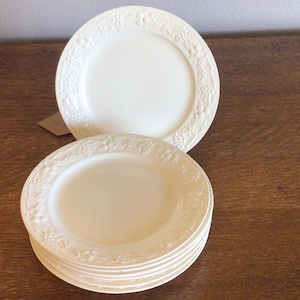 May include: A stack of off-white ceramic plates with a raised grape and leaf design around the rim. One plate is stacked on top of the others. The plates are on a wooden surface.