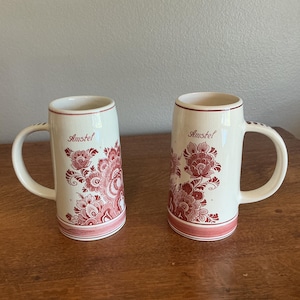 May include: Two off-white ceramic beer mugs featuring a red floral pattern and the word Amstel printed in red script. Each mug has a cylindrical body, a single side handle, and a red band around the base, showcasing a consistent design.