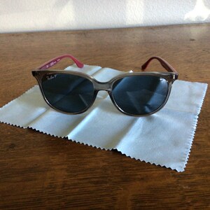 Puede incluir: Gafas de sol con montura y lentes oscuras, sobre un pa&ntilde;o de limpieza blanco. Las patillas son de color rosa rojizo. Las gafas est&aacute;n sobre una superficie de madera.