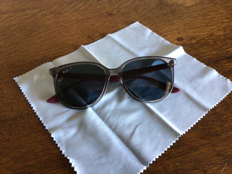 Puede incluir: Gafas de sol con lentes oscuras y montura transl&uacute;cida sobre un pa&ntilde;o de limpieza blanco. Las patillas tienen un acento burdeos. El logotipo de Ray-Ban es visible en la patilla izquierda. Las gafas de sol est&aacute;n sobre una superficie de madera.