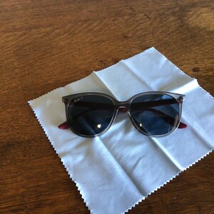 Puede incluir: Gafas de sol grises con lentes oscuras y detalles rojos, sobre un pa&ntilde;o de limpieza blanco. Las gafas tienen una montura rectangular y est&aacute;n sobre una superficie de madera.