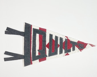 May include: A red, white, and blue pennant with the word "QUILTS" stitched in black. The pennant has a diamond pattern and is made of fabric.