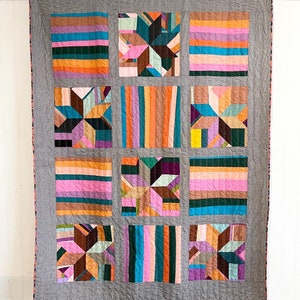 May include: A gray quilt with a patchwork design featuring geometric shapes in various colors, including pink, orange, blue, green, and purple.