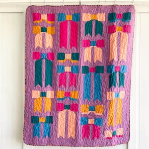 May include: A purple quilt with a repeating pattern of colorful bows. The bows are made of pink, teal, blue, and yellow fabric.